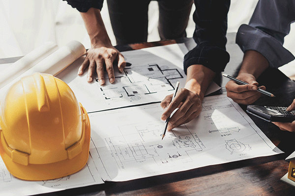 A man’s white hands are visible on a set of design plans. His left hand is planted further back on a table over top the design sheets, and his right hand is holding a pencil close to the paper. The man is wearing a black shirt with white buttons and the cuff of his left sleeve is rolled up. To the left, a woman’s hands can be seen holding a grey pencil and a silver and black calculator at the edge of the design plans. The woman is wearing a grey shirt with the cuff of one visibly rolled up.