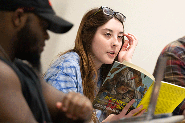 A white female student holds a book and looks to the left in a classroom with a Black male sitting to the right of her.